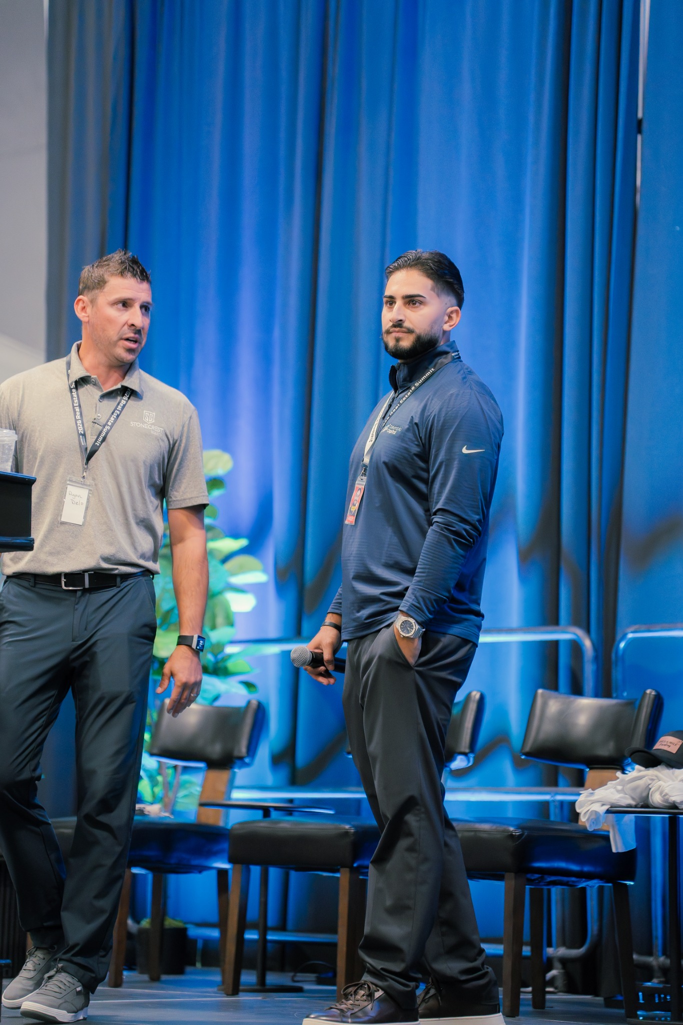 Two presenters preparing together on a blue-lit conference stage.