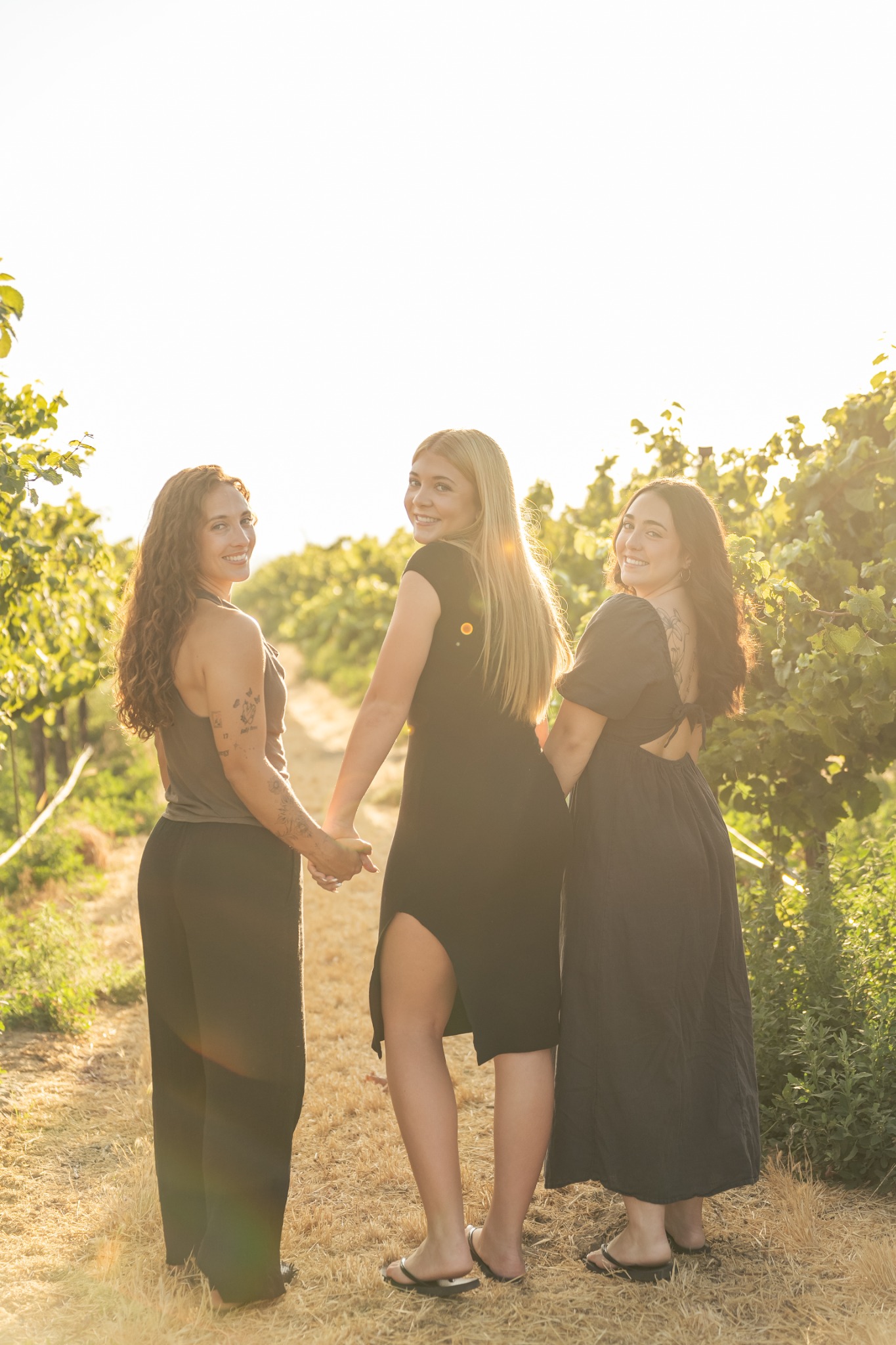 Three sisters holding hands and laughing together between vineyard rows.