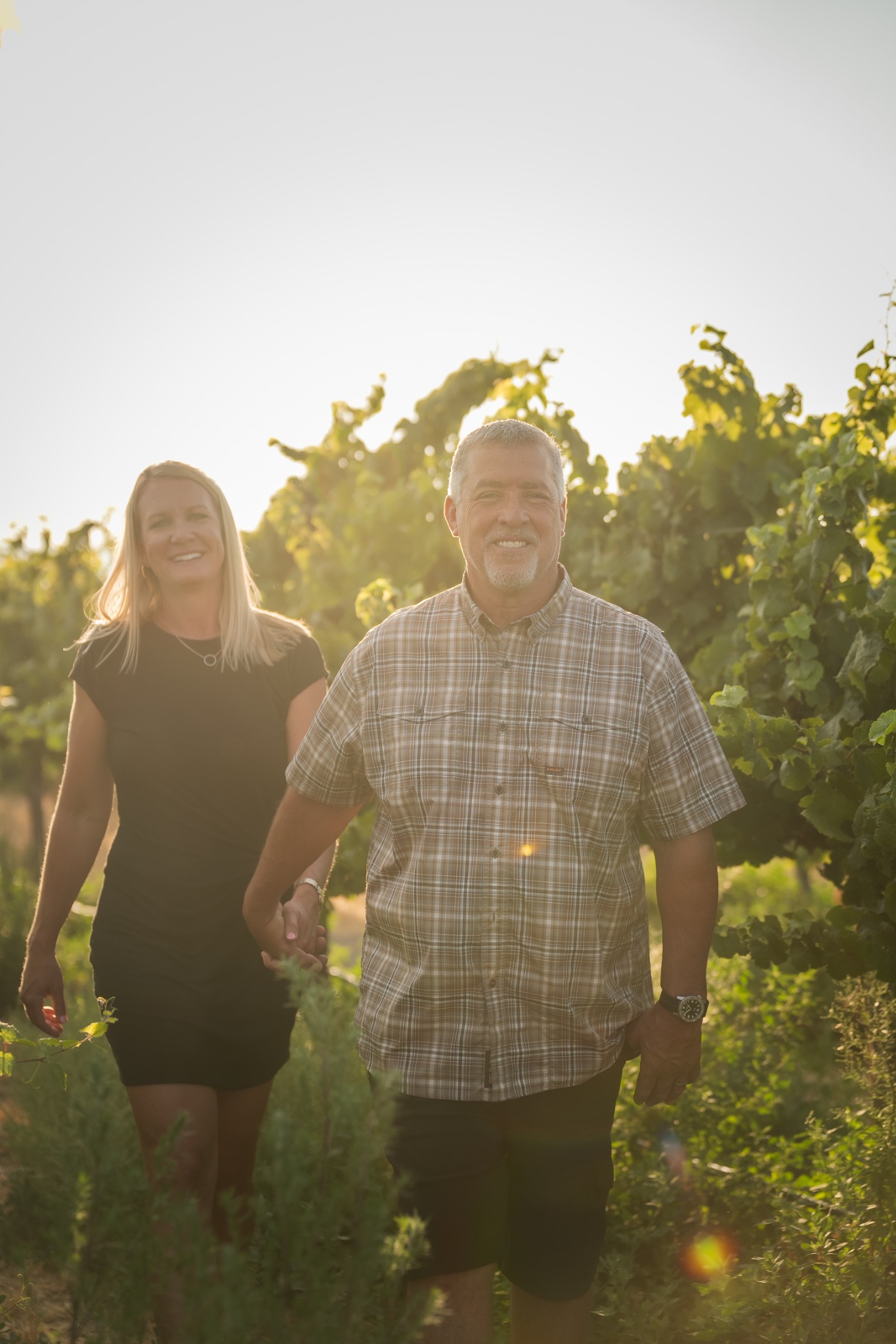 A couple walking hand in hand through a sunlit vineyard.