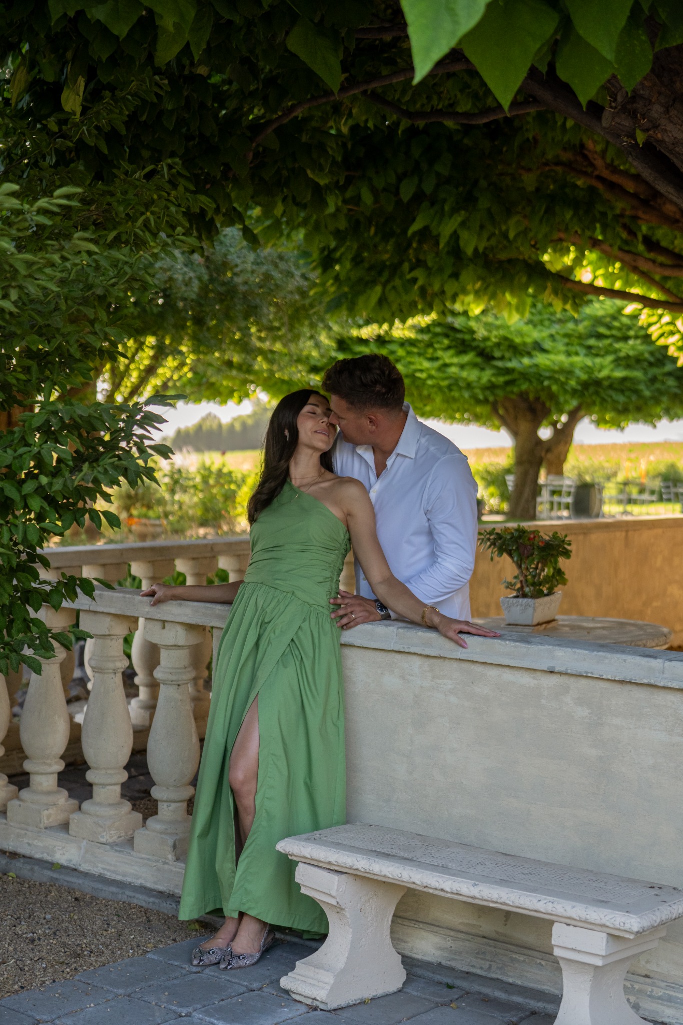 A couple sharing a quiet kiss beneath a canopy of green trees.