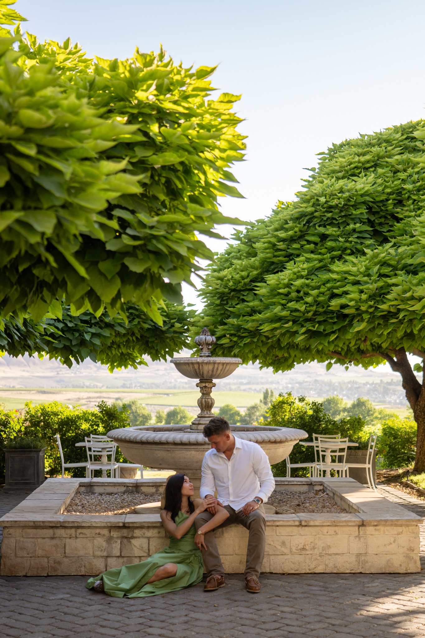 A couple seated beneath sculpted trees with a fountain and vineyard landscape behind them.