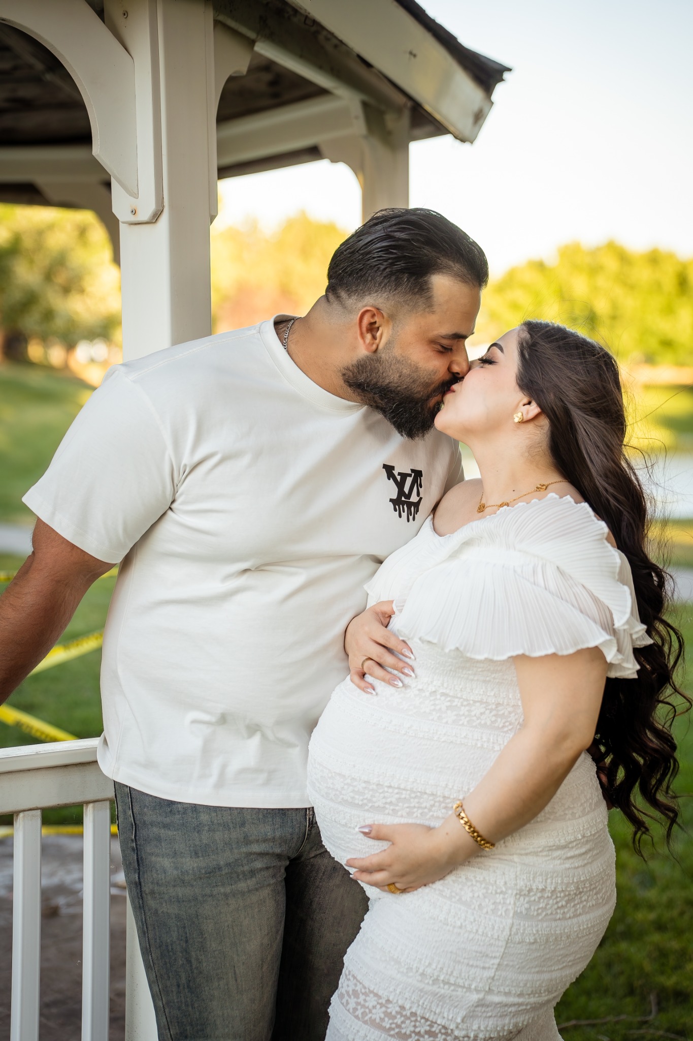 An expecting couple sharing a kiss beside a white gazebo.