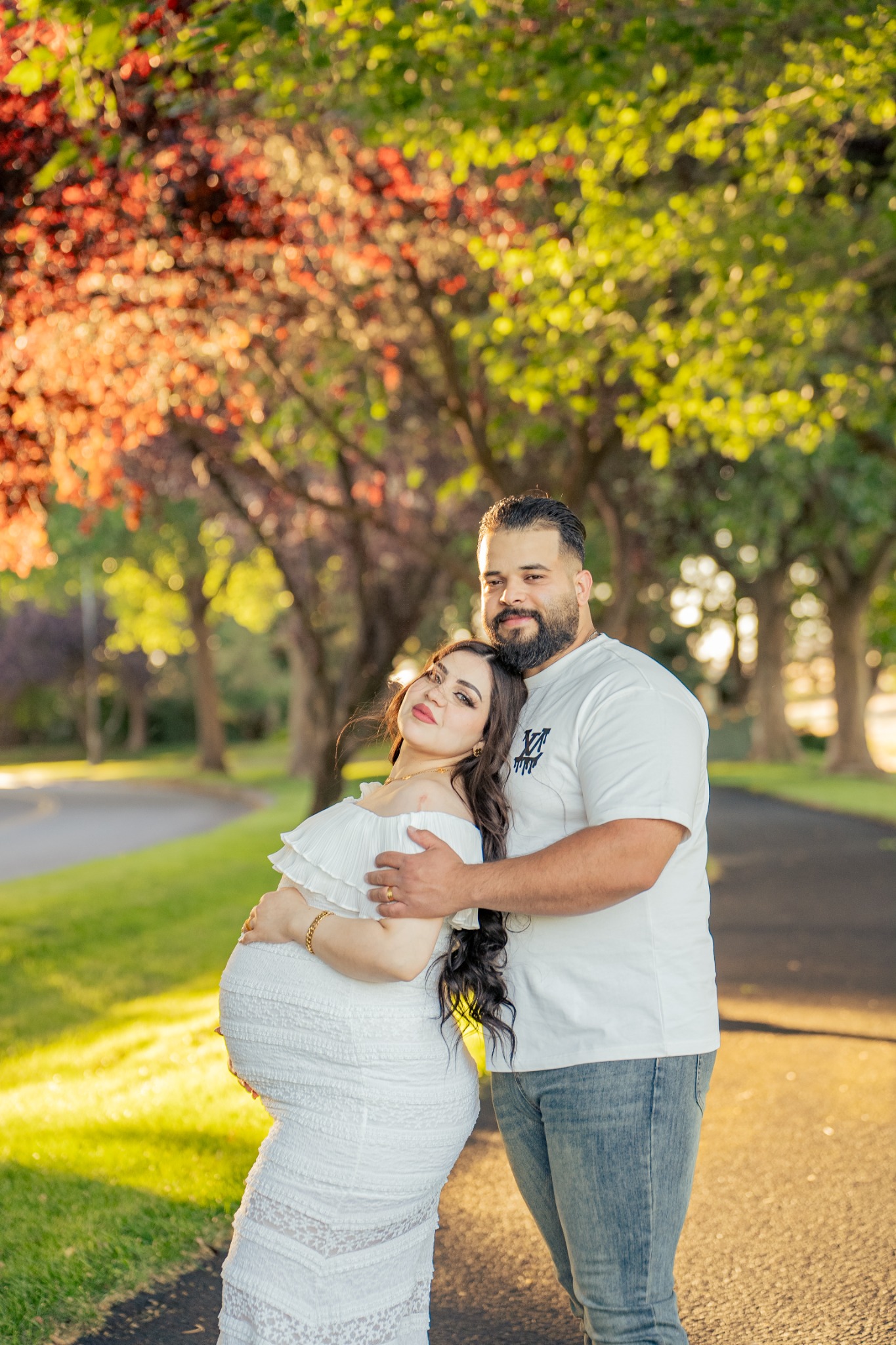 An expecting couple embracing on a tree-lined path in warm evening light.