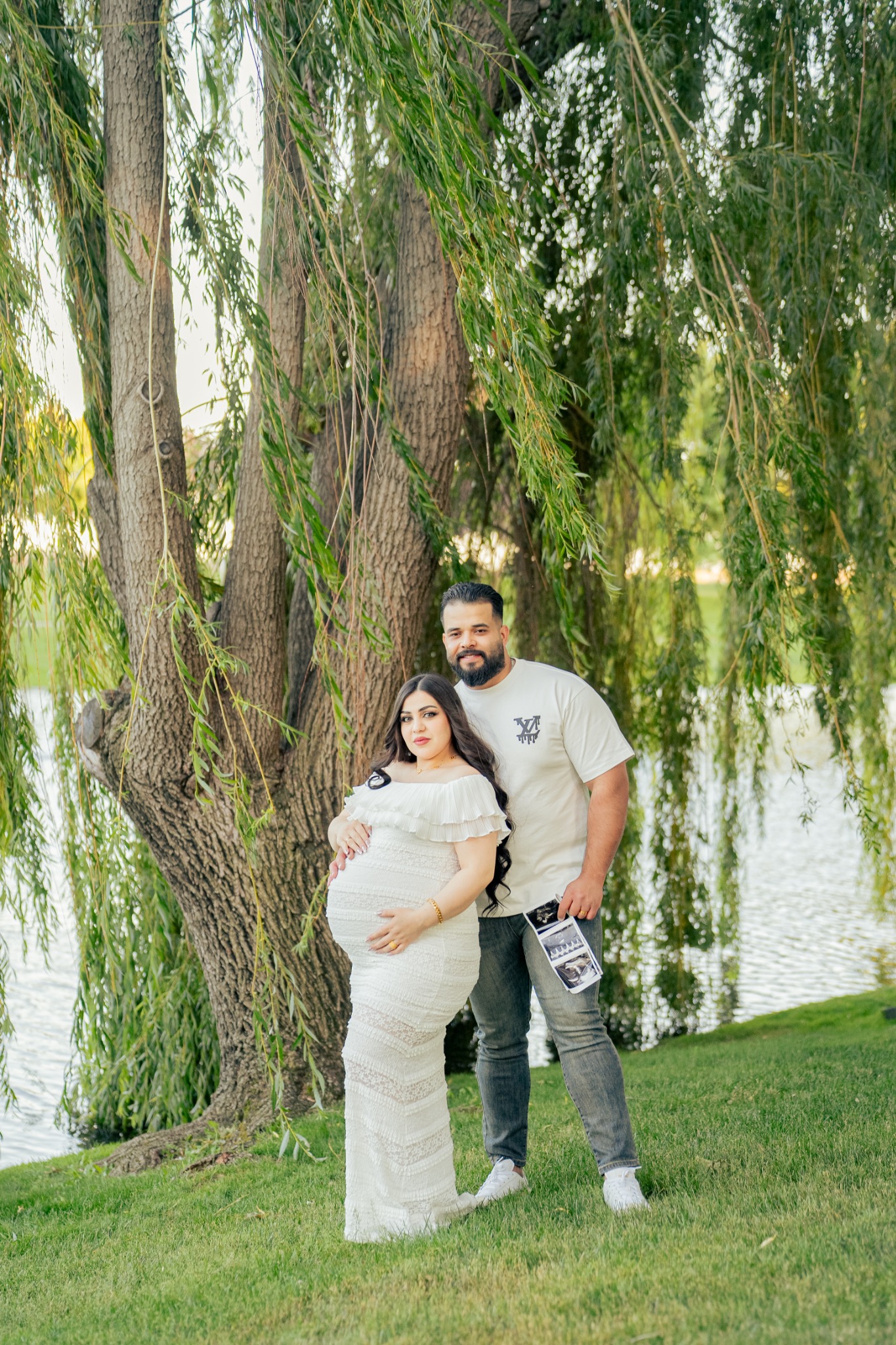 An expecting couple standing beneath willow branches with an ultrasound photograph.