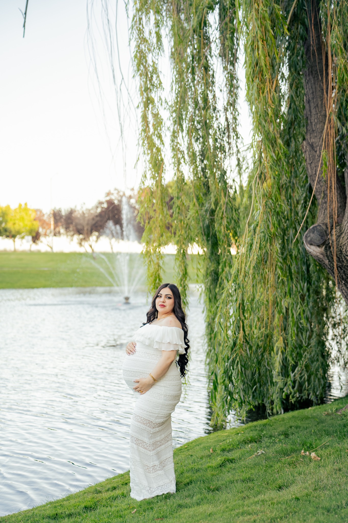 An expecting mother standing beside a pond beneath sweeping willow branches.