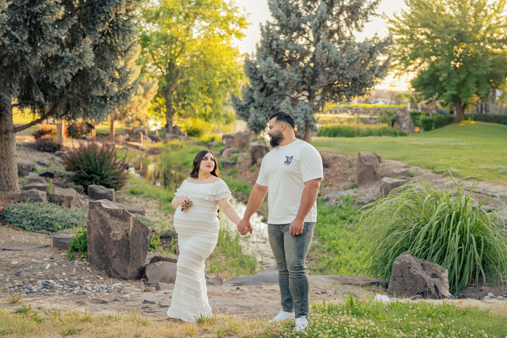 An expecting couple walking hand in hand through a sunlit park.
