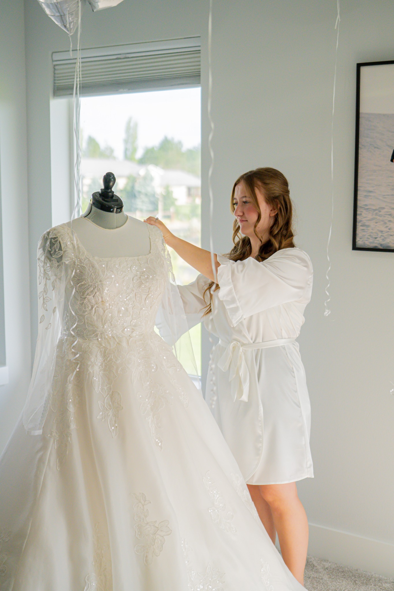 A bride in a robe admiring her wedding dress beside a bright window.